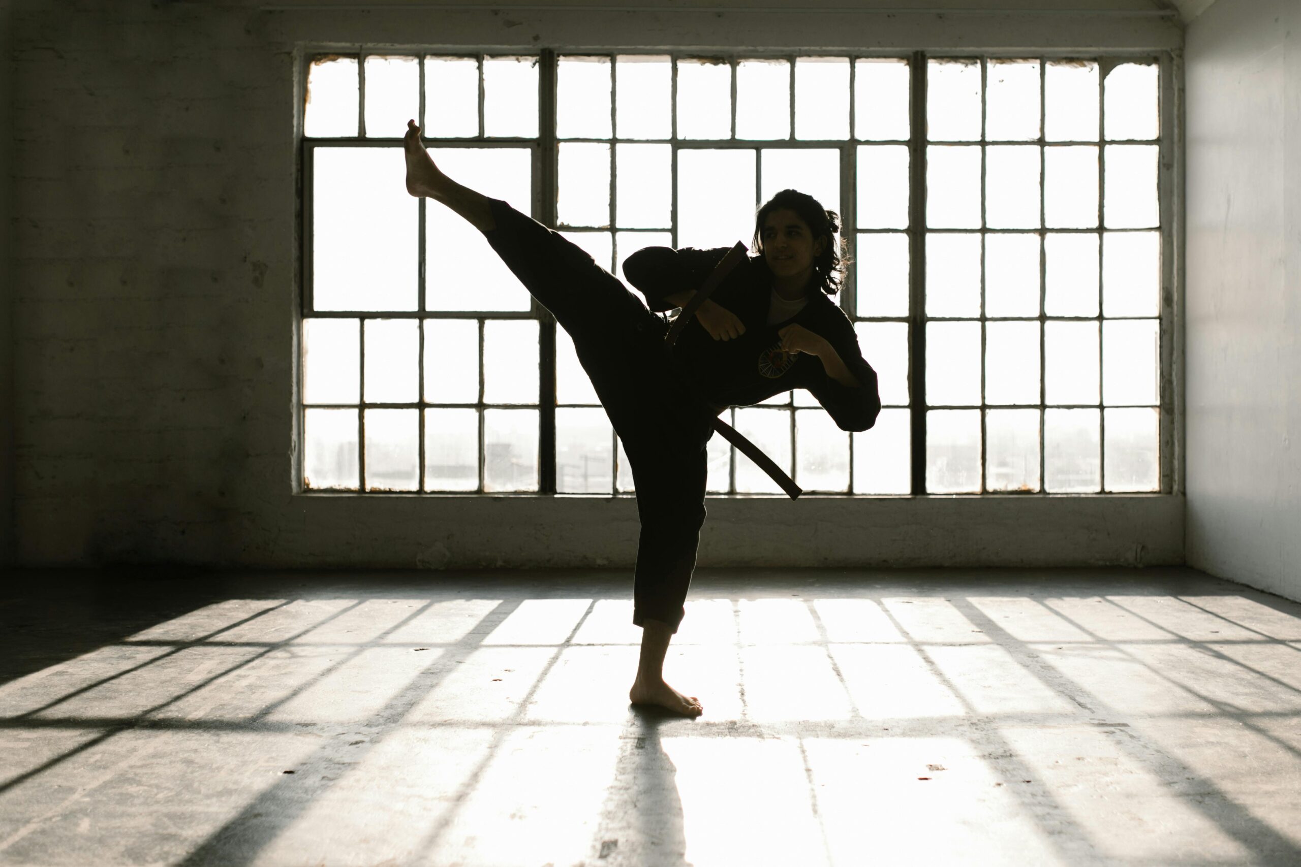 Martial artist performing a high kick in a sunlit room creating dramatic shadows.